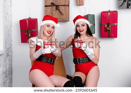 Two beautiful go-go dancers in a red bodysuit with white fur on top, black sandals, New Year's hats and white gloves sit on a background of gifts and drink tea from mugs with a New Year's design