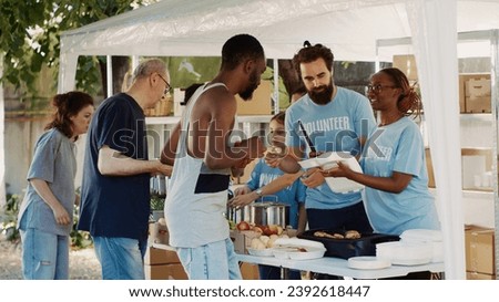 Humanitarian aid team graciously providing free warm meals to the poor needy and less fortunate individuals. Group of volunteers handing out food donations to the homeless people. Tripod shot. Royalty-Free Stock Photo #2392618447