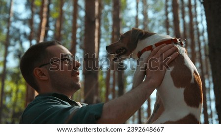 Man holds dog in hands in park. Stock footage. Man happily holds dog in arms in sunny forest. Happy owner keeps dog on background of sun in summer park