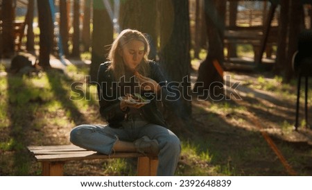 Young woman sitting on bench and eating barbecue and vegetables. Stock footage. Woman with barbecue plate and shining sun through the trees.
