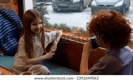 Boy with red curly hair taking photos of a cute long haired girl in a cafe by the window. Stock footage. Children at a table taking pictures.