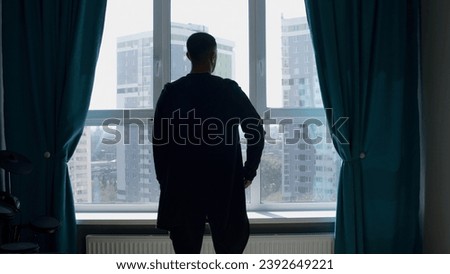 Rear view of a dressed man standing indoors in a living room by the window. Media. Silhouette of a man looking through the window.