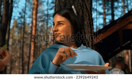 Beautiful brunette woman standing by the tree and eating. Stock footage. Woman taking food from plate, smiling on the background of forest and sky.