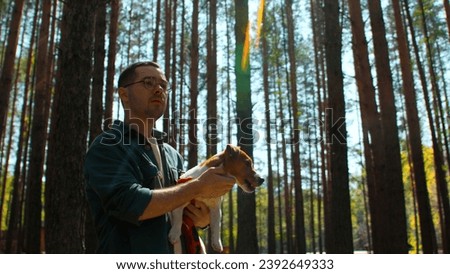 Pensive man in glasses standing among trees in forest with a dog in his hands. Stock footage. Young man in shirt and his puppy.