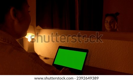 Woman looking at laptop with green screen during night time in front of mirror. Media. Freelancer watching monitor display with chroma key screen, working overtime concept.