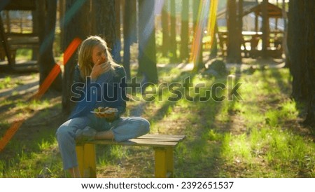 Young woman is sitting on bench and eating barbecue. Stock footage. Beautiful woman is sitting alone and eating barbecue in park in summer. Woman with barbecue plate in nature on sunny summer day