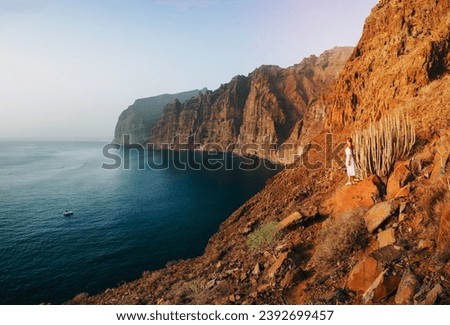 Landscape photo of a woman looking in the distance standing at the edge of big cliffs, the beach and the ocean in the background. Los Gigantes. Incredibly beautiful turquoise water. Tenerife, 