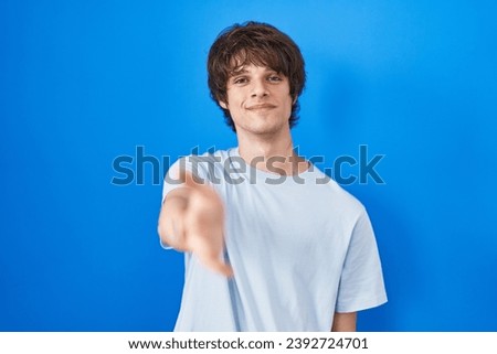 Hispanic young man standing over blue background smiling cheerful offering palm hand giving assistance and acceptance. 