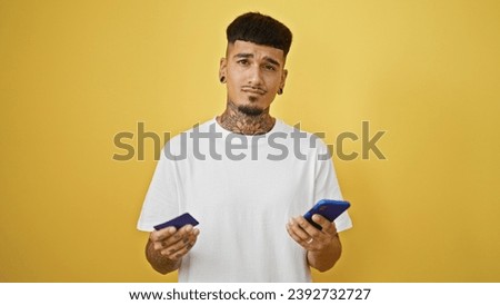 Young, handsome latin man, tattooed and upset, haphazardly uses his credit card and smartphone against isolated yellow wall.