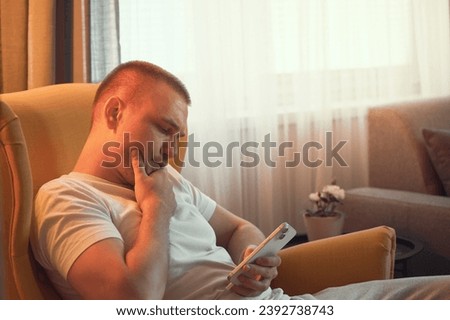 A caucasian man concentrated as he browses social networks on his smartphone, sitting on a yellow cozy chair in his modern house. Man just cruising his phone researching information.