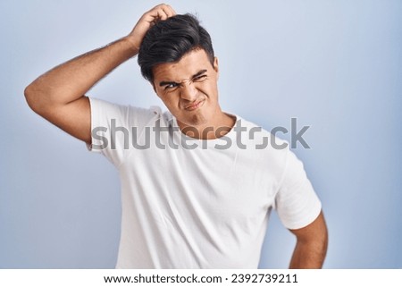 Hispanic man standing over blue background confuse and wondering about question. uncertain with doubt, thinking with hand on head. pensive concept. 