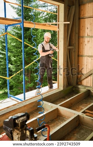 Carpenter constructing wooden framed house. Bearded man worker cladding facade of house, fastening with screwdriver, wearing work overalls and helmet. Concept of modern eco-friendly construction.