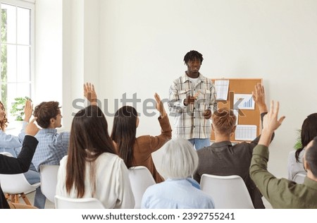 Group of diverse people raise their hands to ask question or vote during business meeting in office. Young African American male coach is speaking in front of people sitting on chairs.