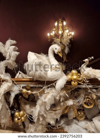 Holiday decoration white swan, golden balls, white feathers on fence with burgundy wall background with wall lamp. White and gold Christmas ornaments and decor.