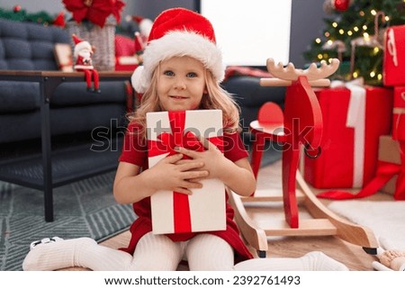 Adorable blonde girl hugging gift sitting on floor by christmas tree at home