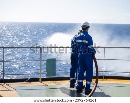 Seamen during fire emergency training drill, on board a merchant cargo ship, wearing fire fighting equipment and helmets. Rigged fire hose for jet spray, with emergency fire pump. Royalty-Free Stock Photo #2392764261