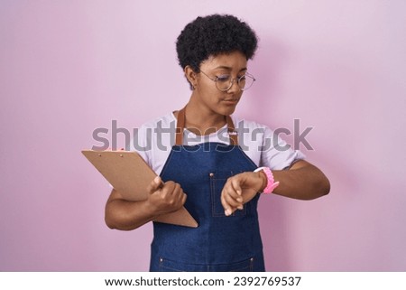 Young african american woman wearing professional waitress apron holding clipboard checking the time on wrist watch, relaxed and confident 