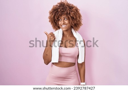Young hispanic woman with curly hair wearing sportswear and towel smiling with happy face looking and pointing to the side with thumb up. 