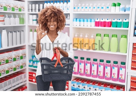 Young hispanic woman with curly hair shopping at pharmacy drugstore holding basket doing ok sign with fingers, smiling friendly gesturing excellent symbol 