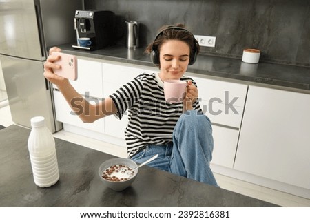 Portrait of young woman, content maker for social media, taking selfie while drinking tea and listening music in headphones, posing for photo, using smartphone to take pictures in the kitchen.