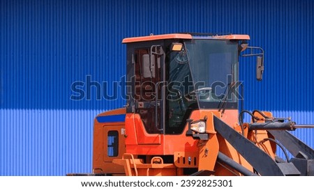 Cab control room of orange hydraulic excavator in front of blue warehouse building wall in construction site, close up with copy space
