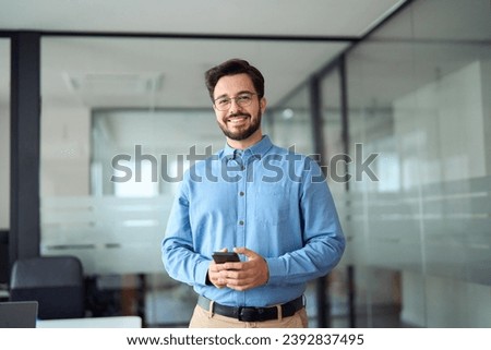 Smiling busy professional latin business man standing in office holding mobile cellphone. Young happy businessman employee using smartphone looking at camera using cell phone tech at work. Portrait. Royalty-Free Stock Photo #2392837495