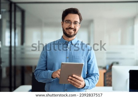Smiling busy young latin business man manager using tablet computer, happy hispanic businessman executive looking at camera holding tab working standing in office. Portrait. Royalty-Free Stock Photo #2392837517