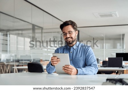 Smiling busy young latin business man manager using tablet computer, happy hispanic businessman executive looking at tab device analyzing finance trading market data working sitting at office desk.