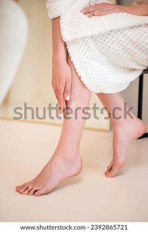 Vertical shot of woman in soft robe sitting in bathroom, applying moisturising cream after shaving legs on skin. Female in bathrobe using pampering products for skincare. Beauty procedure at home Royalty-Free Stock Photo #2392865721