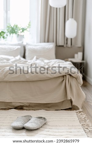 Selective focus on home slippers on floor carpet near comfort bed with pillows, blanket and bedding linen. Cozy bedroom with scandinavian interior in light tones. Hygge style in room