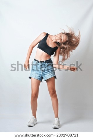 Beautiful young model in a casual black top and jeans shorts dancing and shaking head with long hair on a grey background