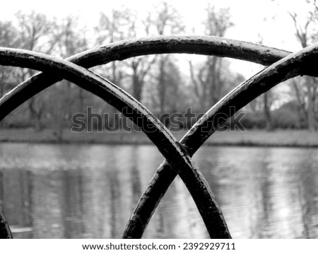A black gloss painted metal fence by the side of the lake at University Park, Nottingham, UK.  View through the railings over the water beyond.  Overlapping bars make an A shape.  March 2015. Royalty-Free Stock Photo #2392929711