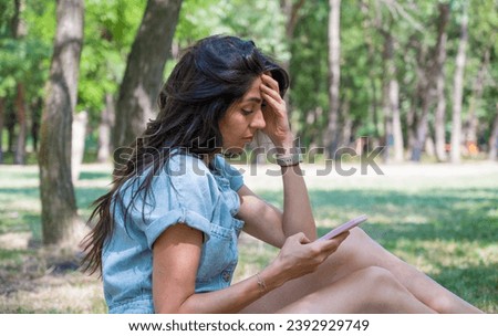 Beautiful young sad woman sitting in the summer park looking her phone  Royalty-Free Stock Photo #2392929749
