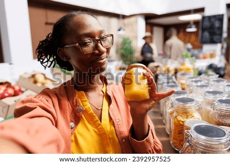 During video call, african american lady with glasses clutches a glass jar of honey. At eco friendly store, black woman uses phone for vlogging and leverages technology for internet marketing. Royalty-Free Stock Photo #2392957525