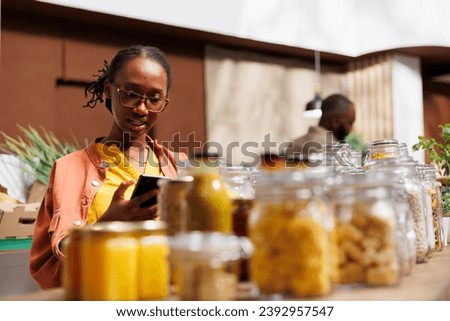 African American woman on her smartphone, surrounded by eco friendly products and reusable jars. Image shows a young black lady using her mobile device to compare products and prices.