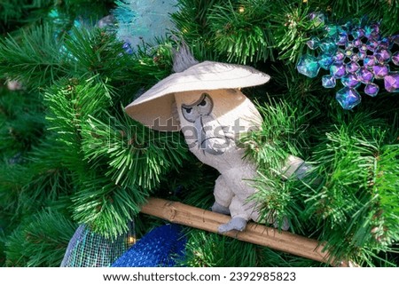 A small white bird character with a mean and angry face wearing a wide brim sun hat. The animal's hair is poking out through the center of the hat. The Christmas figurine is hanging in a green tree.