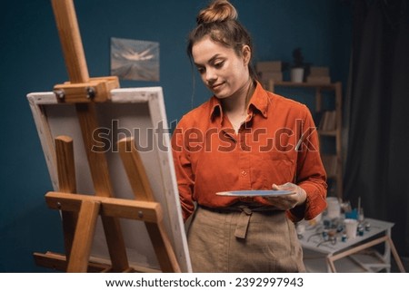 Young beautiful painter woman standing in modern workshop painting picture on canvas with paints, enjoy creative hobby. Education, and art-class concept