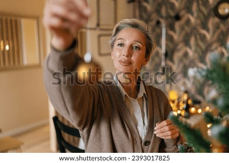 Portrait of happy middle-aged woman holding gold ball for decoration Christmas tree at home, closeup, bokeh lights, selective focus. Concept of home festive atmosphere.