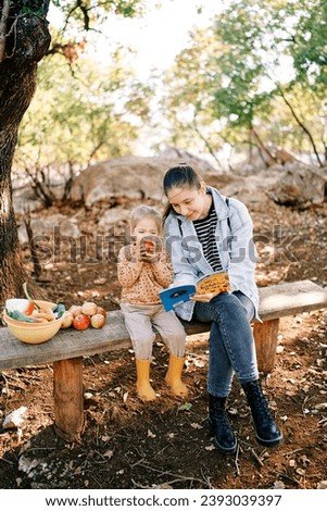 Mom reads a book to a little girl gnawing an apple while sitting on a park bench