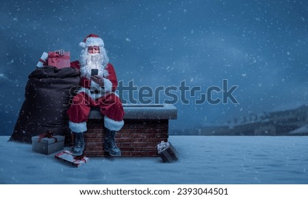 Santa Claus delivering gifts on Christmas Eve, he is sitting on a chimney and connecting online with his smartphone Royalty-Free Stock Photo #2393044501