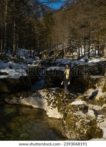 Cheerful woman standing on a large mossy stone in the middle of a mountain river. Attractive young tourist lady went for a winter walk by the pristine river that flows from heart of alpine mountains. Royalty-Free Stock Photo #2393068191