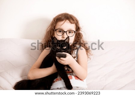 Cute curly little girl kid child in a eyeglasses watching TV with kitty cat best friend at home with remote control in a hands