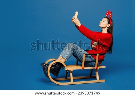 Full body merry young Latin woman wear red Christmas sweater decorative fun deer horns on head sledding do selfie shot on mobile cell phone isolated on plain blue background. Happy New Year concept
