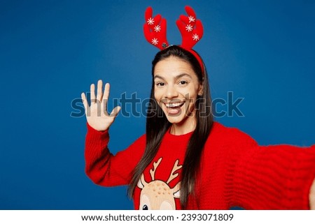 Close up young Latin woman wear red Christmas sweater decorative fun deer horns on head posing do selfie shot on mobile cell phone waving hand isolated on plain blue background. Happy New Year concept