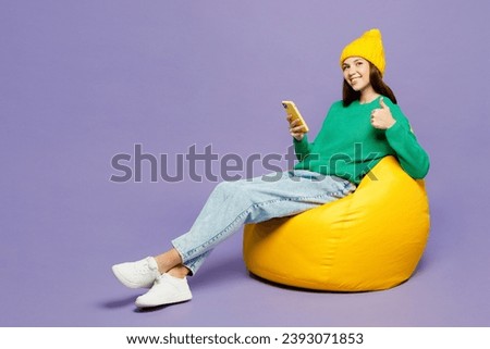 Full body young woman wear green sweater yellow hat casual clothes sit in bag chair hold in hand use mobile cell phone show thumb up isolated on plain pastel light purple background. Lifestyle concept