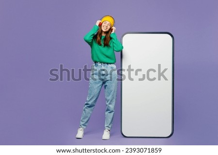Full body young woman she wear green sweater yellow hat casual clothes big huge blank screen mobile cell phone smartphone with area listen to music in headphones isolated on plain purple background