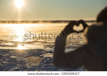 Woman making heart with hands outdoors in a frosty sunset, closeup. Focus on ice floes. High quality photo