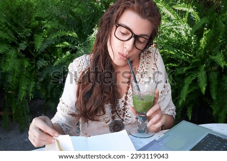 A young woman in glasses sits outdoors, working on a laptop and drinking a green juice. She studies and communicates digitally, surrounded by nature. A perfect picture of remote work and education.