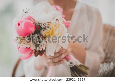 The bride in a white wedding dress holds a wedding bouquet. The bouquet consists of white, pink, peonies and pink berries. The bride has a wedding ring on her hand.