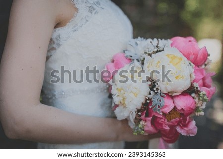 The bride in a white wedding dress holds a wedding bouquet. The bouquet consists of white, pink, peonies and pink berries. The bride has a wedding ring on her hand.